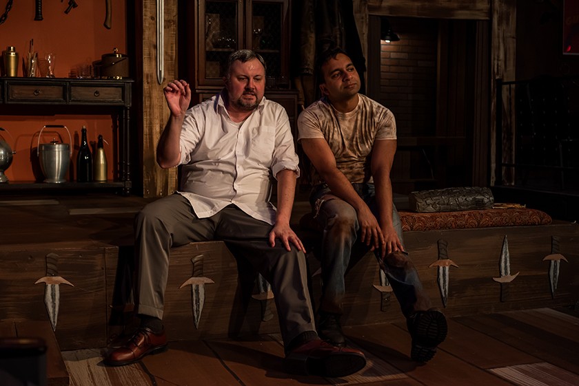 Two men sit side-by-side on a theater stage. The man on the white is white, with short brown hair and a goatee, a dirty white collared shirt, and gray pants. The man on the right has dark brown skin, short black hair, and is wearing a dirty white T-shirt, and dark pants