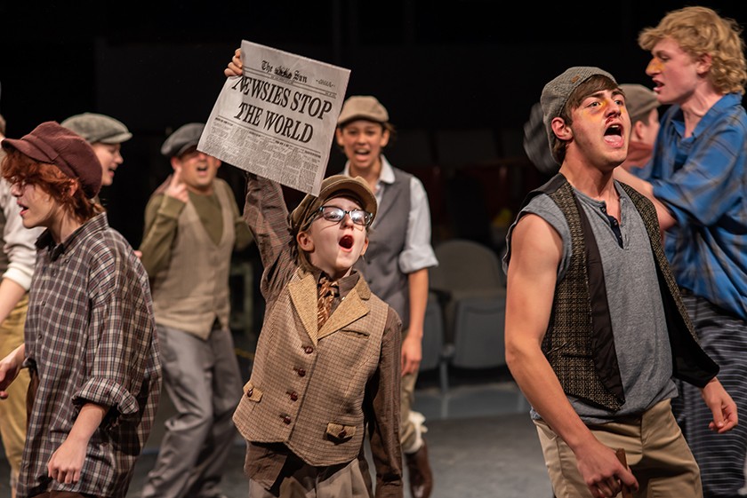A crowd of young actors on stage wearing old fashioned clothing. The young boy in the center of the photo is white and wears glasses, a newsboy cap, a long sleeved brown shirt, best, and tie, and is holding up the front of a newspaper that reads "Newsies stop the world"
