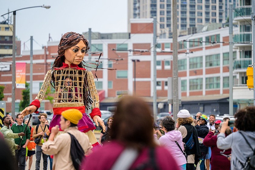 A giant 12-foot-tall puppet of a young Syrian girl stands amid a crowd of humans in a city scene. A person controlling the puppet is visible through the puppet's torso.