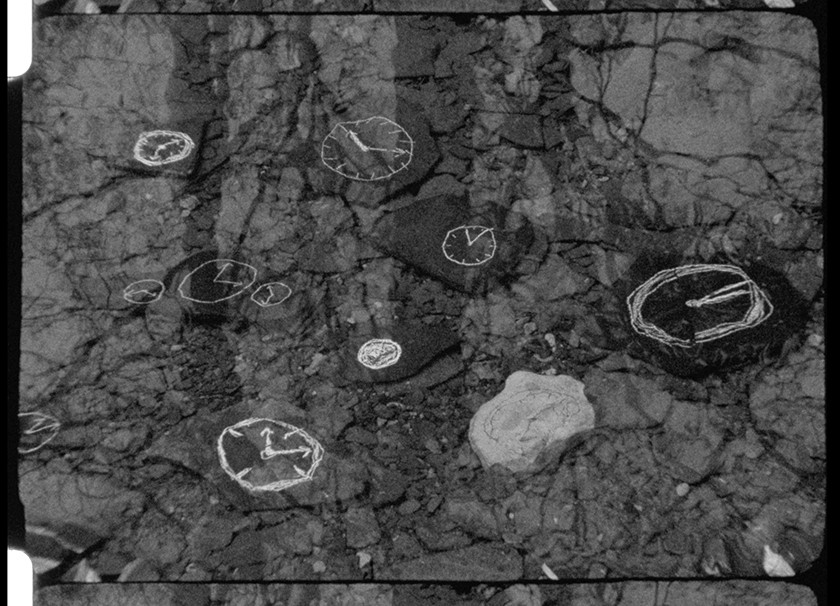 A black-and-white film still showing white sketches of clock faces on a dark rough exterior surface