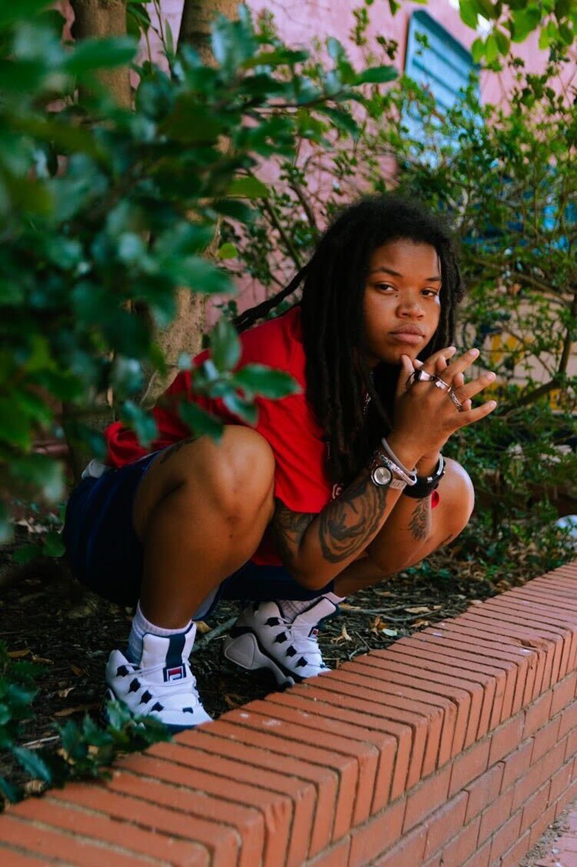 A young Black person with long locs and wearing a red T-shirt, shorts, and sneakers, kneels down on top of a brick flower bed, outside of a building