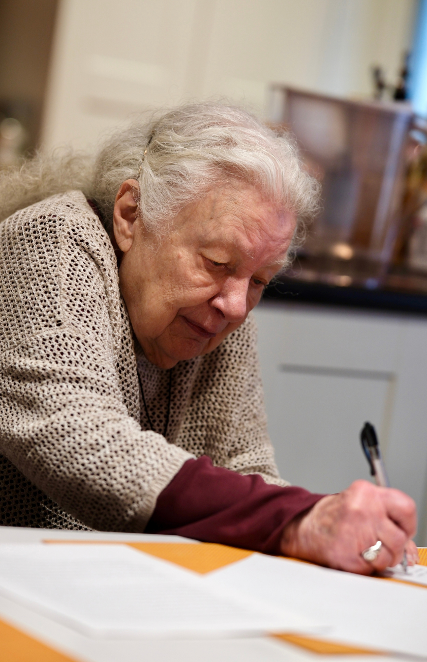 An older woman with white hair writing on paper with a pen.