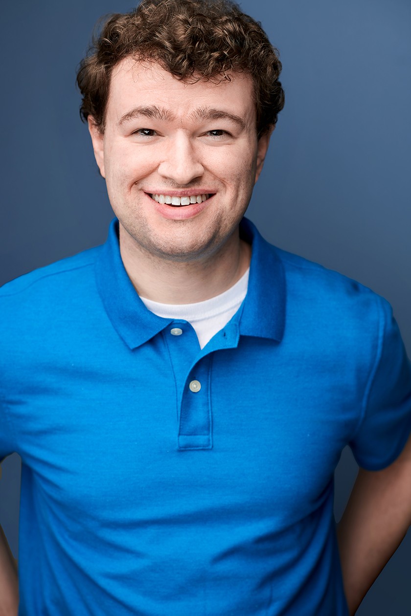 A smiling headshot of a person with short curly brown hair wearing a white t-shirt underneath a bright blue polo shirt
