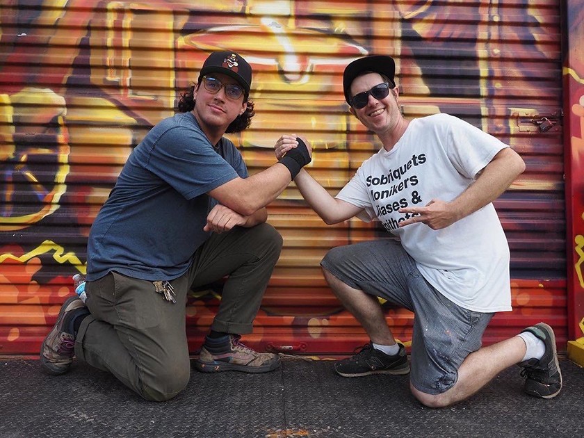 Two people kneel down and grip hands. Both are smiling. The person on the left is wearing a blue T-shirt, Army-green pants, a black baseball cap, and glasses. The one on the right has on a white T-shirt, gray shorts, dark sunglasses, and a black baseball cap. The one on the right is flashing a peace sign. Both are posed in front of a colorful mural.