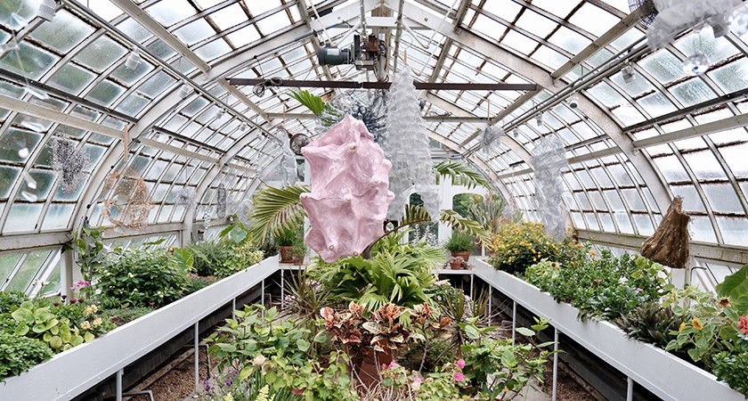 A glass greenhouse filled with various green plants and flowers. In the center, there is a large pink abstract sculpture surrounded by lush foliage. The greenhouse has a curved, windowed roof letting in natural light.