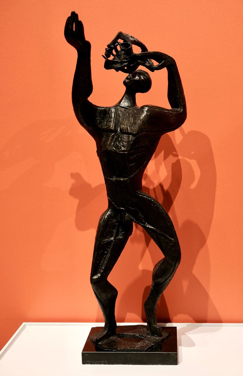 A dark abstract sculpture of a standing human figure with exaggerated limbs and raised arms, posed dynamically against an orange background, displayed on a white pedestal.