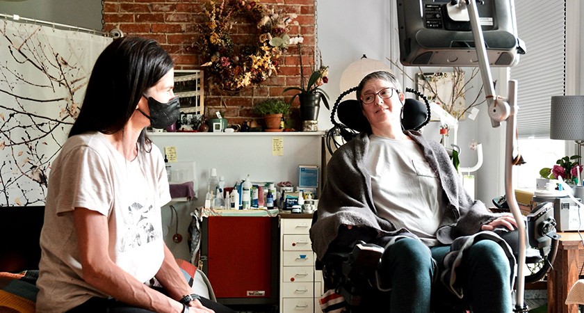 Two women sit indoors; one wears a mask and sits on a chair, while the other sits in a motorized wheelchair with a headrest. Medical supplies and home decor are visible in the background.