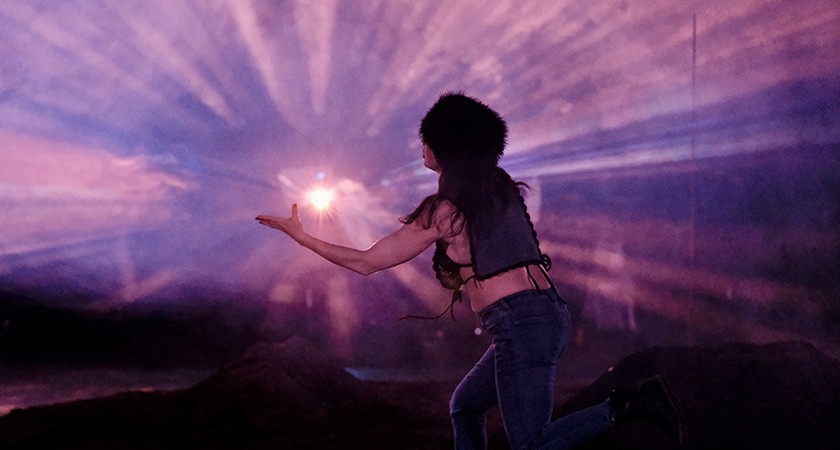 A person wearing a furry hat and jeans kneels with one arm extended toward a glowing light, surrounded by dramatic purple and pink rays in a dark, misty environment.