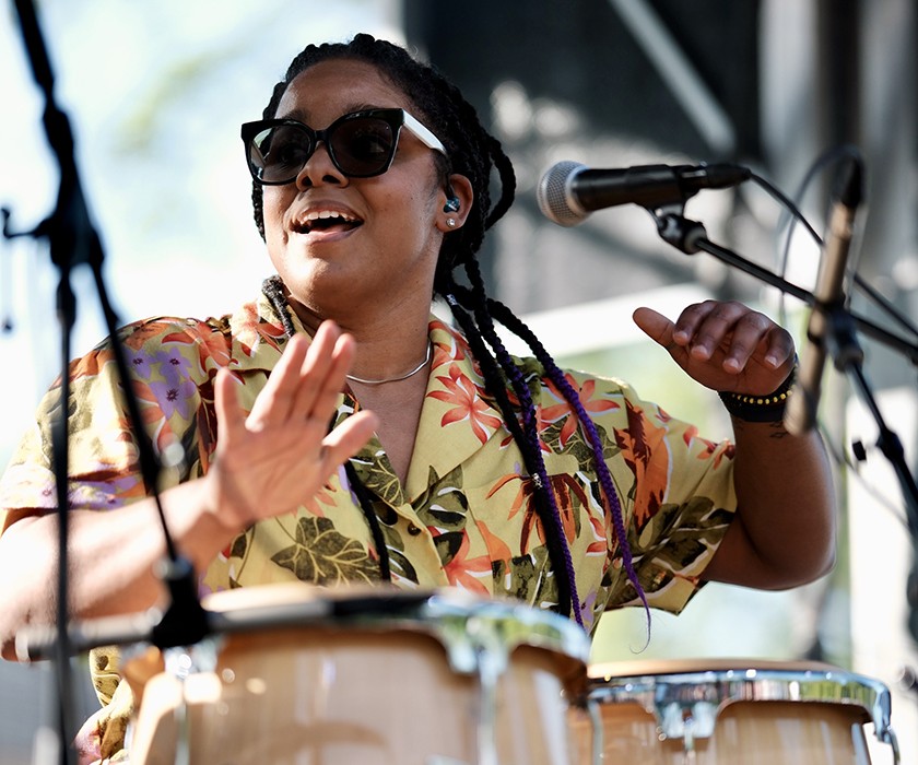 A musician wearing sunglasses and a floral shirt plays conga drums and smiles while performing onstage in daylight. A microphone is positioned near their mouth.