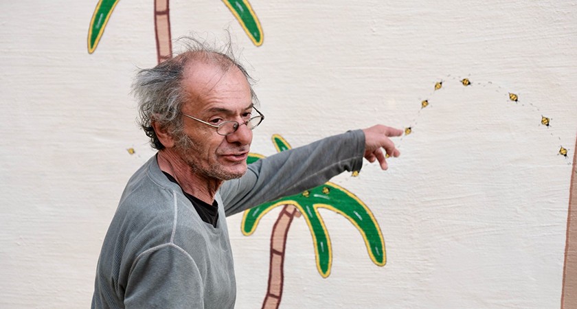 A man with gray hair and glasses points at a mural on a wall featuring palm trees and a dotted line with small painted bumble bees along it.