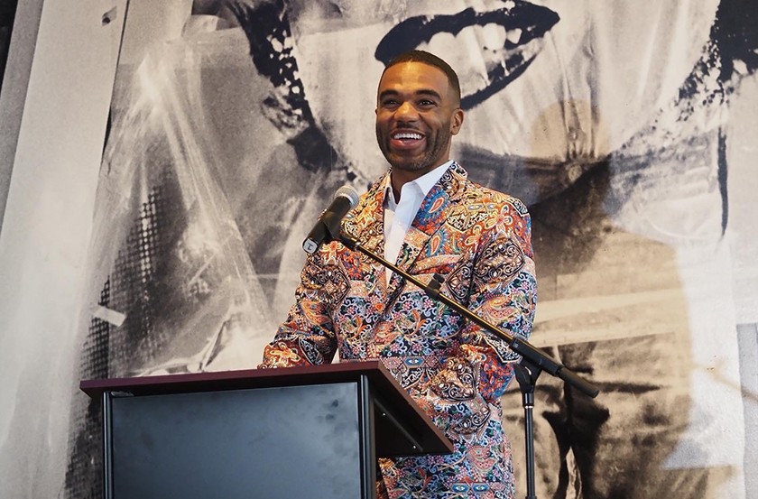A Black man in a multi-colored suit stands smiling behind a podium and in front of a black-and-white silkscreened image of Marilyn Monroe and Andy Warhol