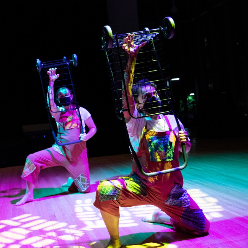 Two performers in overalls kneel on a theater floor while holding up metal wheeled carts above their heads