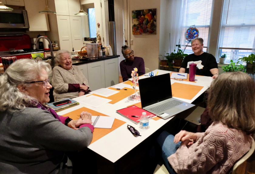 Five women sit around a table with laptops and documents, participating in a meeting.