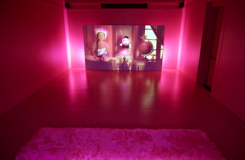 A pink-lit room with a screen showing a child and a doll, and a fluffy bench in front.
