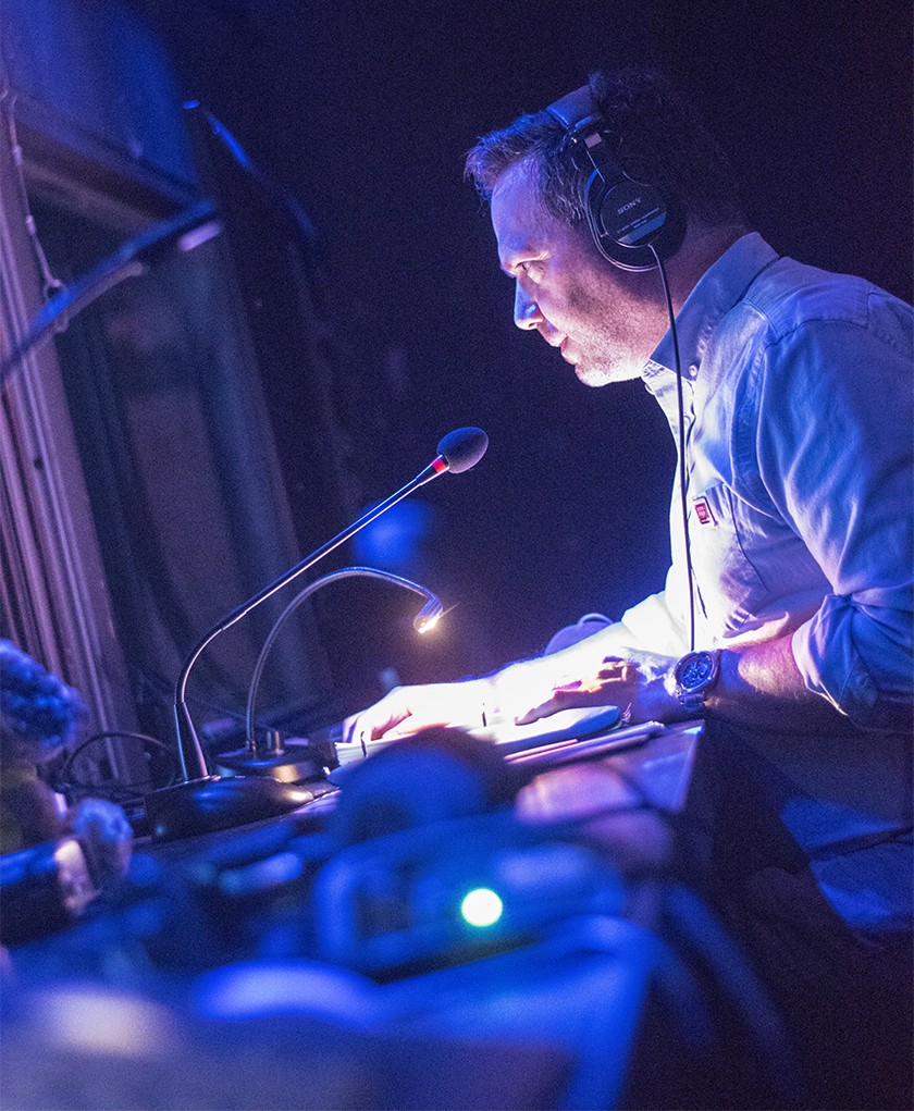 Photo of Nathan Ruggles. Inside a theater tech booth, from a vantage point low above a long desk running away along its length, he sits in profile, leaning toward a mic sitting on the cluttered surface, gazing intently out through the window at left. A middle-aged male wearing headphones and a sky blue oxford cuffed at the elbow, a small table lamp reflects striking white light up into the light pink skin of his face, casting deep shadows in what otherwise is a space dimly lit in blue and lavender.