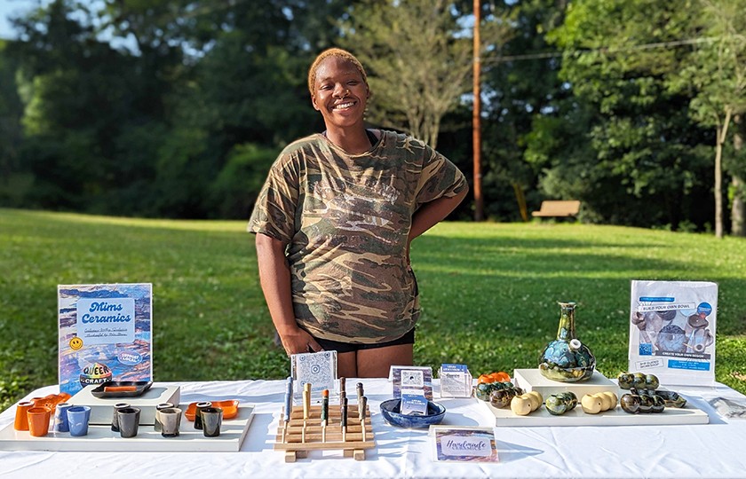 A Black person with short light hair wearing a camo-patterned shirt and black shorts stands smiling in front of a table featuring an assortment of handmade ceramic objects