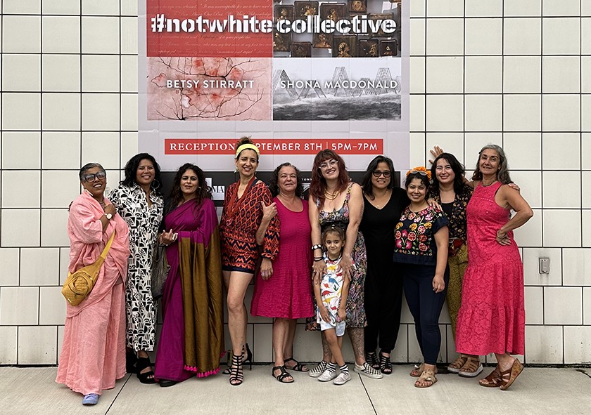 10 Black and Brown women and one child stand in a row in front of a sign that says #notwhite collective