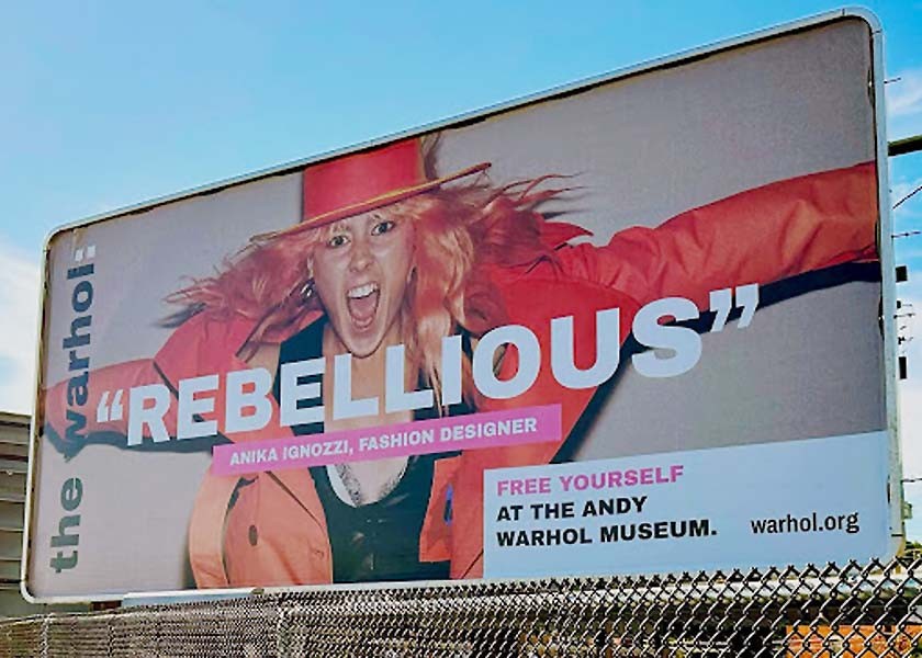 A billboard showing a photograph of a woman wearing a brightly colored pink jacket and hat. She's happy yelling. The word "rebellious" is written on top of the photo