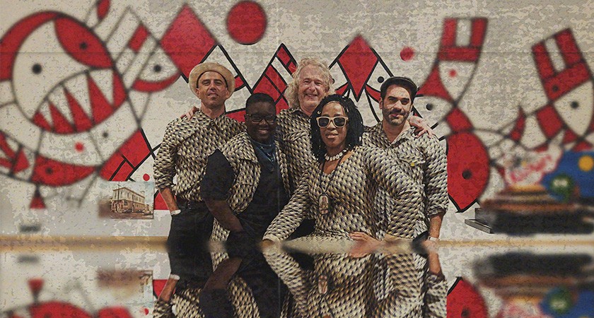 Five people in matching checkered suits pose closely together in front of a red, white, and black mural