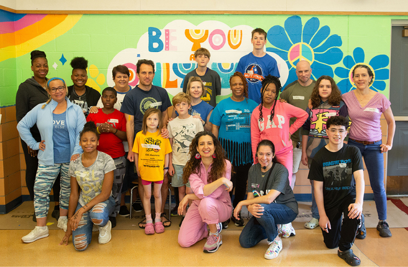 A diverse group of people pose and smile together in front of a colorful mural