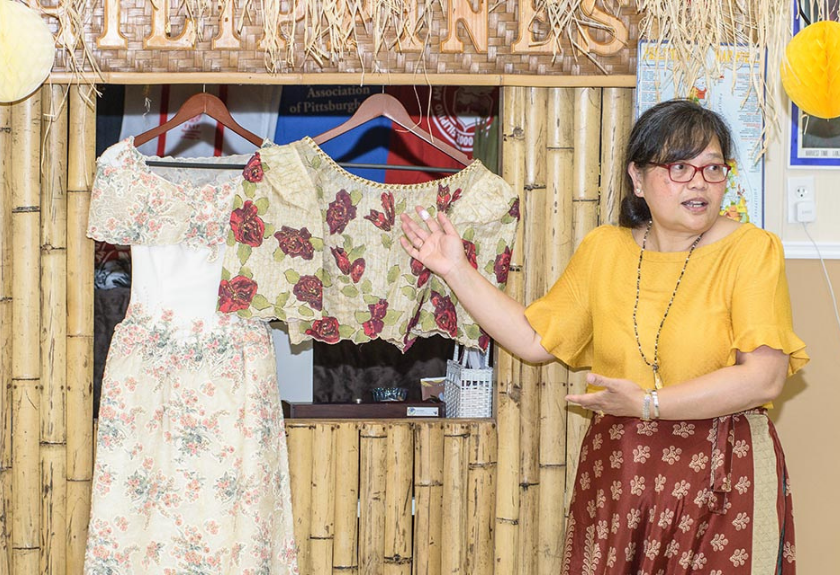 Woman presenting traditional dresses displayed on hangers in front of a bamboo backdrop.