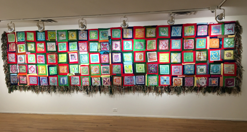 Colorful quilt squares displayed in rows on a wall, with fringe along the bottom edge