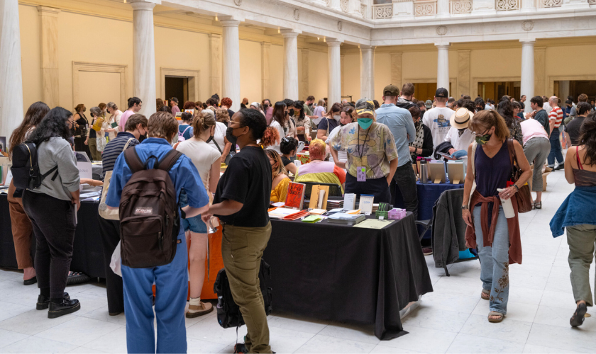 A large group of people browse tables with books and other items at an indoor event in a bright, spacious hall with white columns and marble floors. Some attendees wear masks and backpacks.