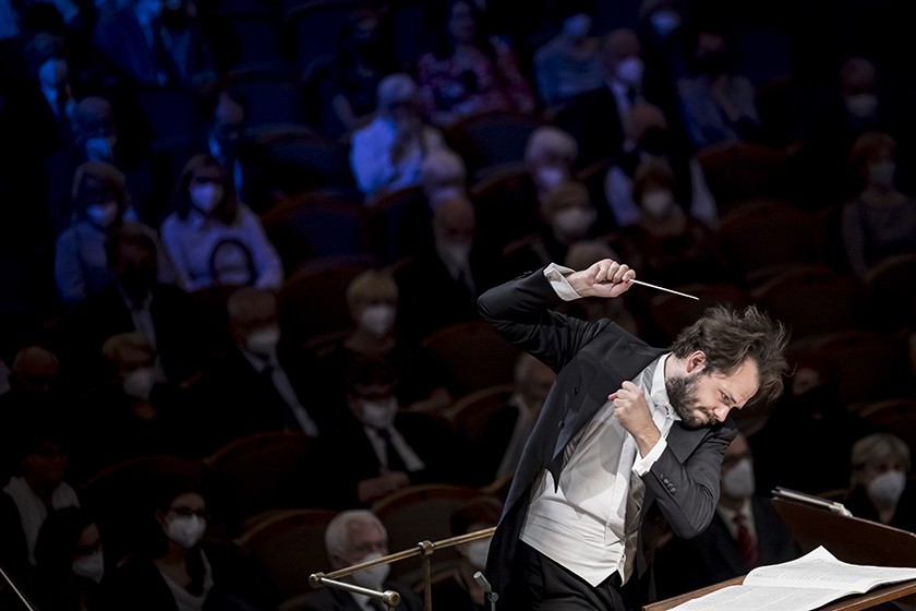 A man with a dark brown beard and mustache, and dark brown short hair wearing a white shirt, black suit jacket, and black slacks is shown conducting a symphony in front of a group of masked audience members in the background