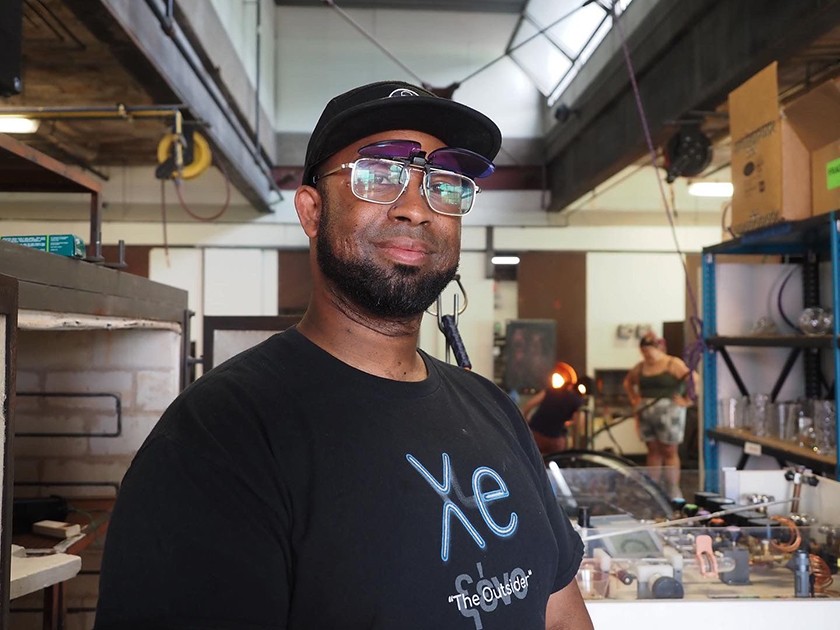 A Black man in a black T-shirt, black baseball cap, and glasses smiles while standing inside a studio space