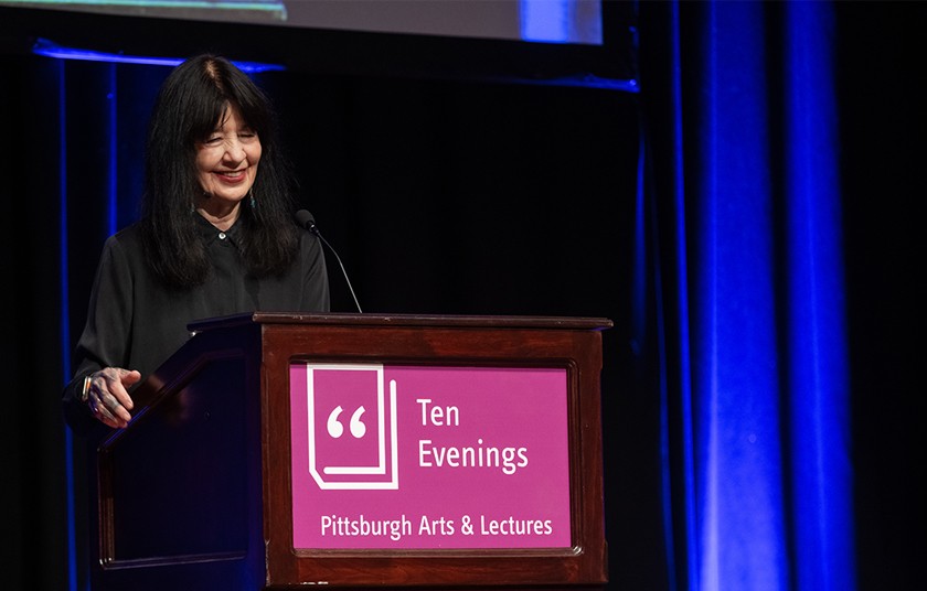 Woman with dark hair stands smiling behind a podium that reads, "Ten Evenings, Pittsburgh Arts & Lectures"