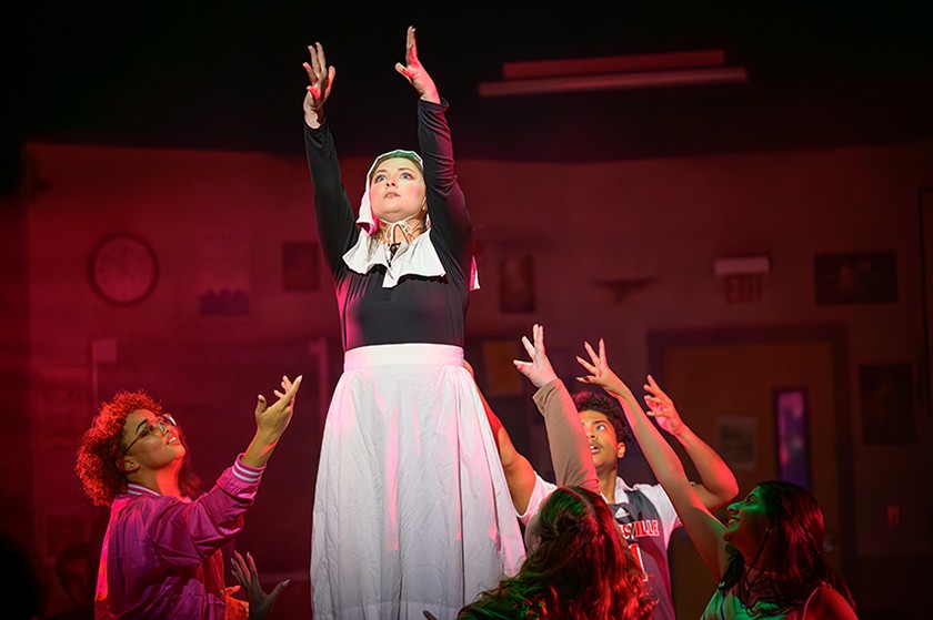 A cast of actors stand on stage raising their arms in the air. In the middle is an actress dressed as a nun