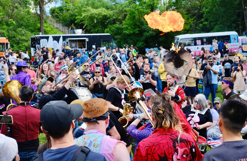 Outdoor brass band performance with a crowd; fire bursts from a tuba in the center.
