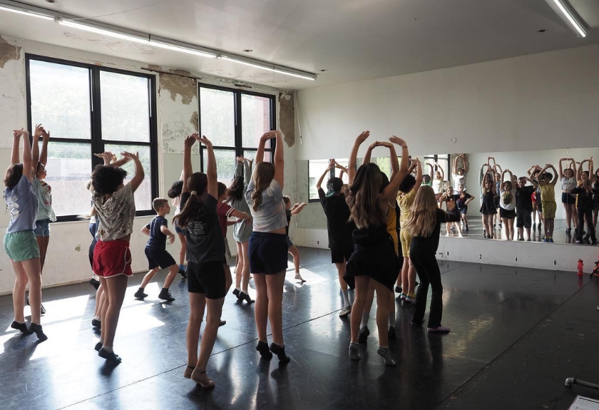 A group of dance students practice in a studio with arms raised above their heads.