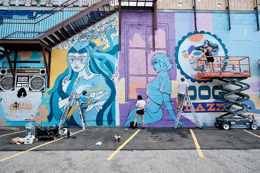 Three artists use lifts and ladders to paint a colorful mural with large blue and purple female figures and vibrant patterns on a tall urban wall in a parking lot. Art supplies are scattered on the ground.