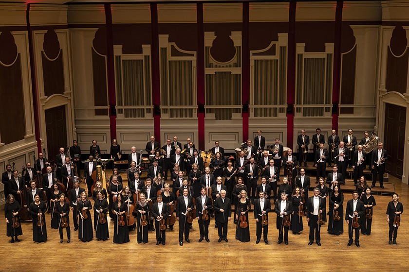 A large group of orchestral musicians wearing black-and-white formal wear and holding various instruments stand on a concert stage