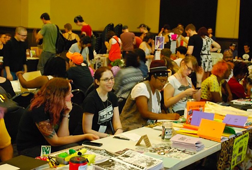A room full of people sitting behind tables displaying a variety of zines