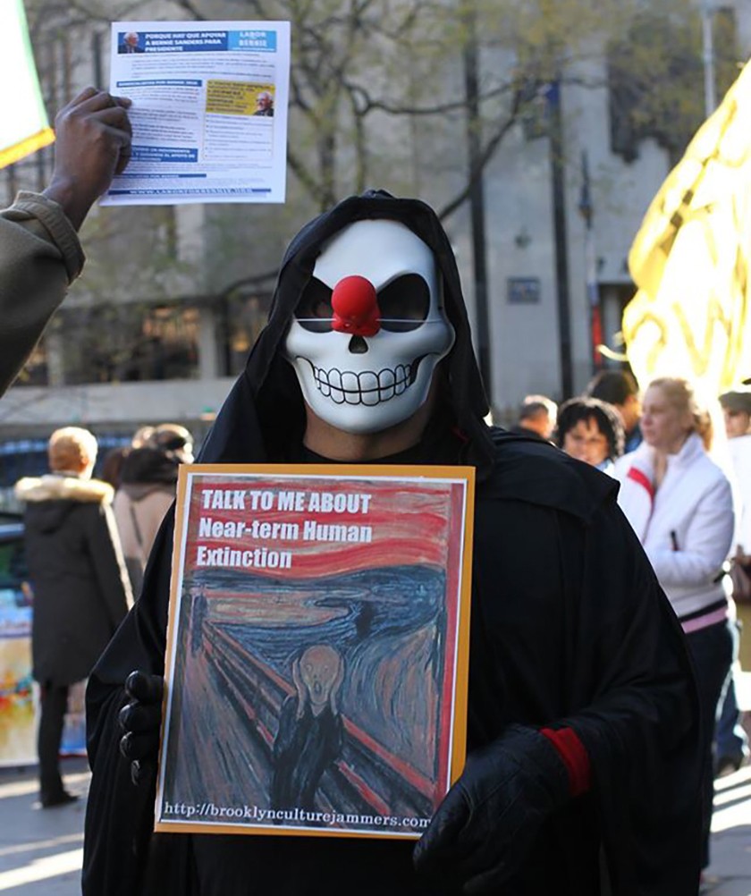 A person dressed as the grim reaper holds up a framed sign featuring the words "Talk to me about near-term human extinction" written on top of the famous Scream painting