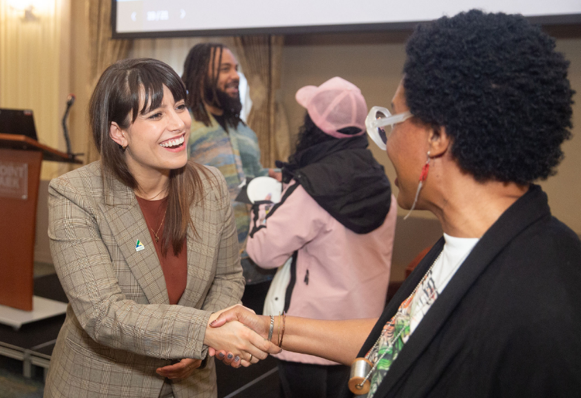 Two women shaking hands and smiling at an indoor event, with two others in the background.