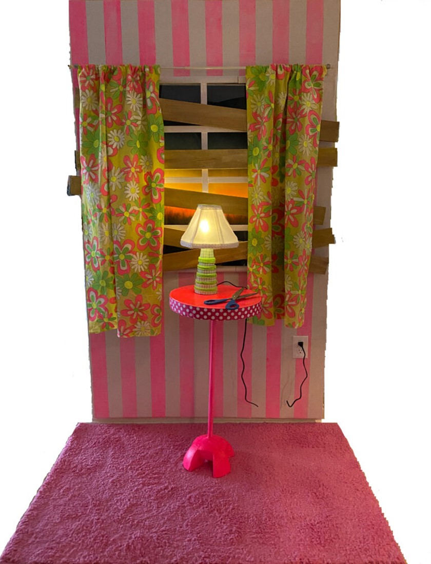 Art installation showing a lamp and scissors sitting on a small pink table, which is placed on a fuzzy pink rug. In the background is a pink-and-white striped wall, a boarded up window covered in retro floral printed curtains
