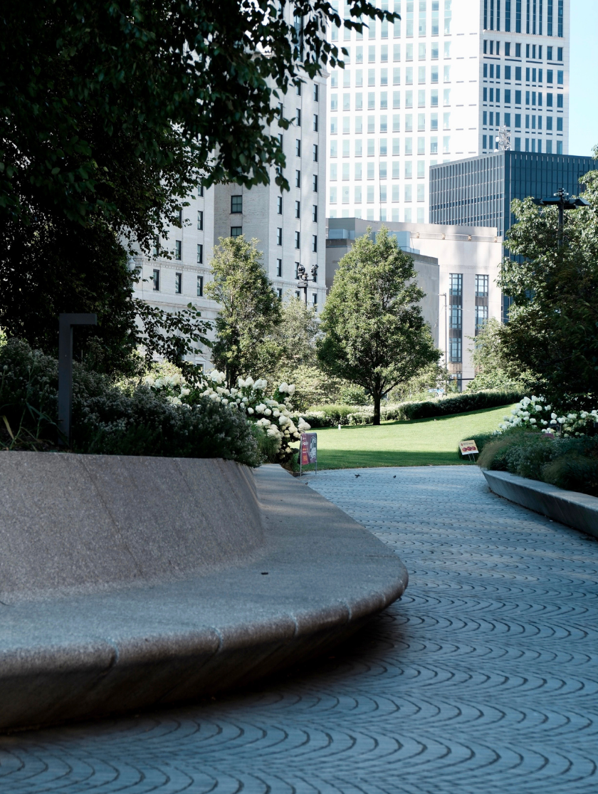 Curved stone bench by a winding path in an urban park with trees and tall buildings behind.