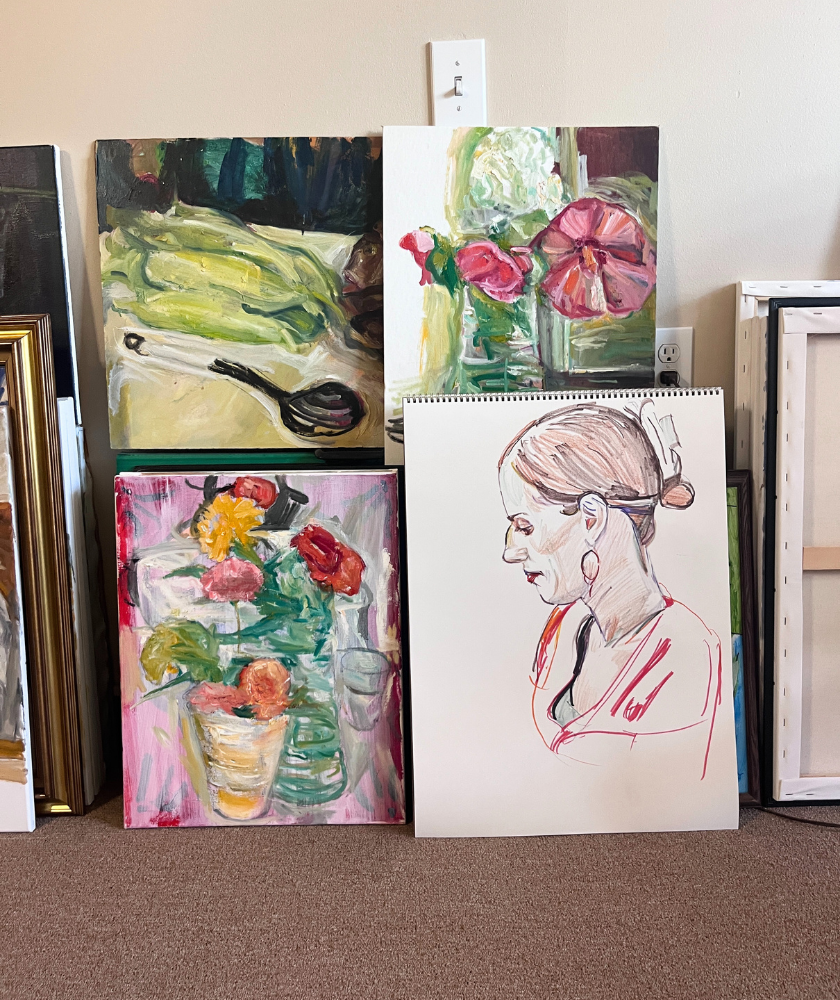 Four colorful paintings stand against a wall: three still lifes featuring flowers and glassware, and one sketch of a woman in profile with her hair up and earrings. The artworks are propped on a carpeted floor.