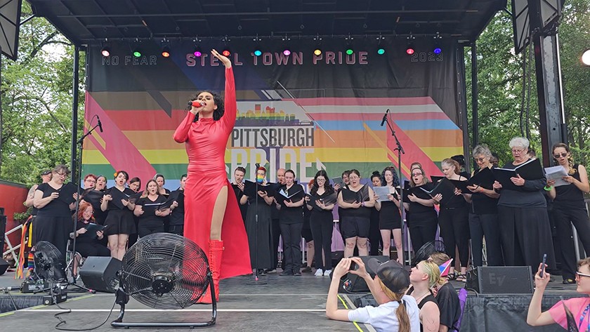 A person in a long red dress sings while holding a microphone. A choir of people wearing black clothing stand behind them on stage in front of an LGBTQ rainbow flag