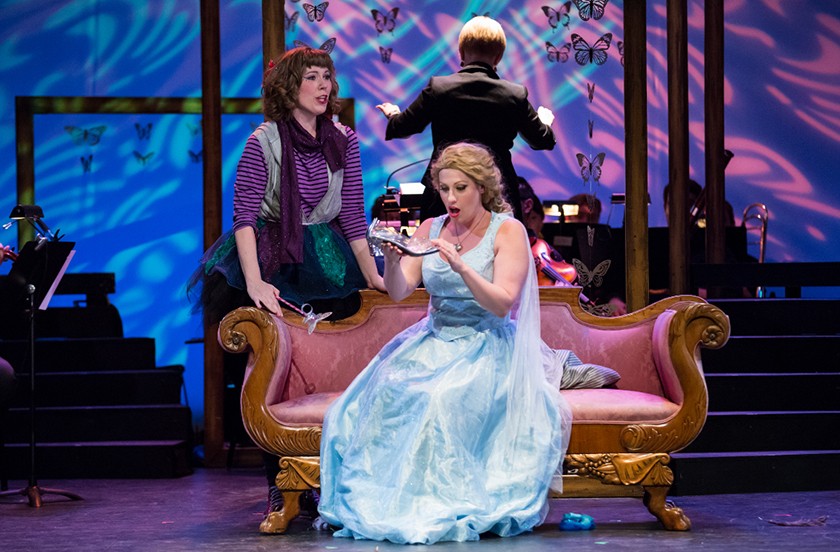 Three costumed actors on stage. An actress in the front of the stage is sitting on a couch wearing a light blue dress holding a slipper