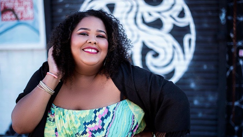 Smiling woman with shoulder-length curly black hair, red lipstick, gold bangle bracelets, a bright blue-green-purple-and-blue patterned dress, and a black pullover