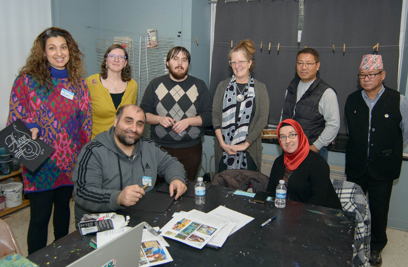 Eight adults pose together in a classroom around a table with art supplies and papers.