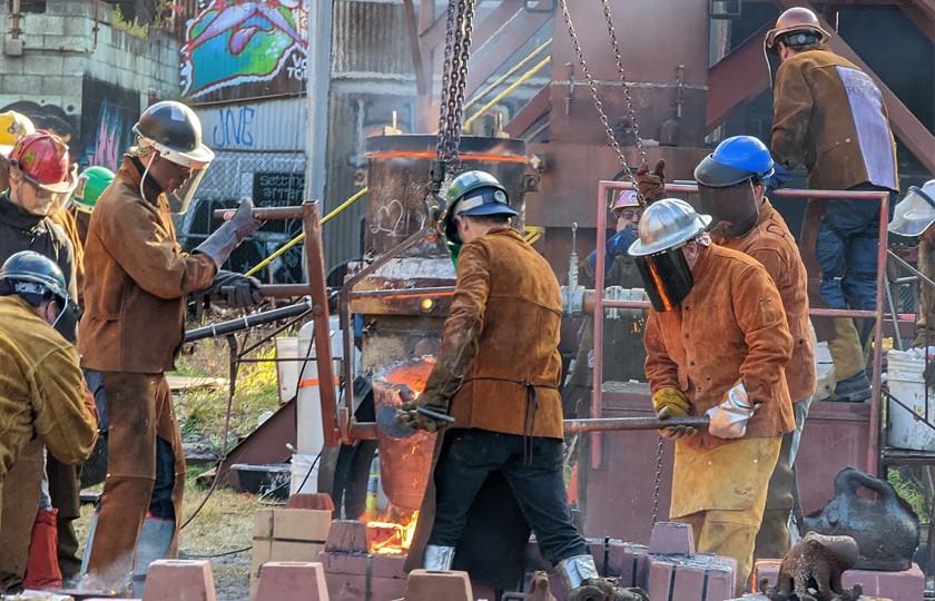 A group of steel workers wearing helmets stand outside pouring hot steel into molds