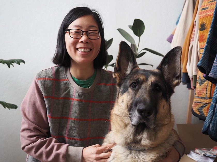 A smiling Asian woman with shoulder-length black hair and sunglasses. She's wearing a purple, gray sweatshirt over a green T-shirt and holding onto a large dog who's staring into the camera. Behind them are a rack of handmade clothing.