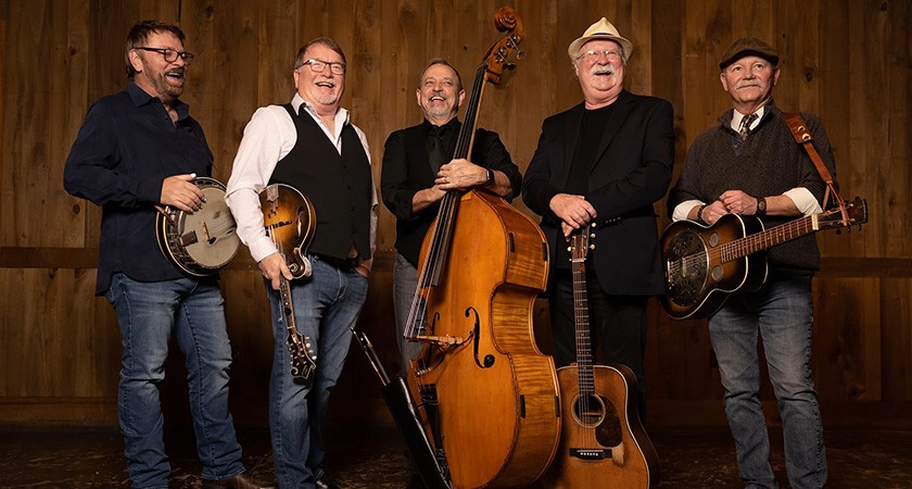 Five smiling older men wearing jeans and dark shirts are shown holding various string instruments