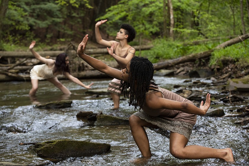Three dancers pose dramatically in a river. They're hands and legs are stretched out. There are large rocks in the river and trees visible in the background