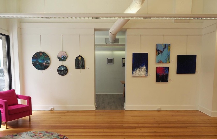 A gallery space showcasing artwork hanging on a wall, wooden floors, a colorful purple sitting chair, and white beams